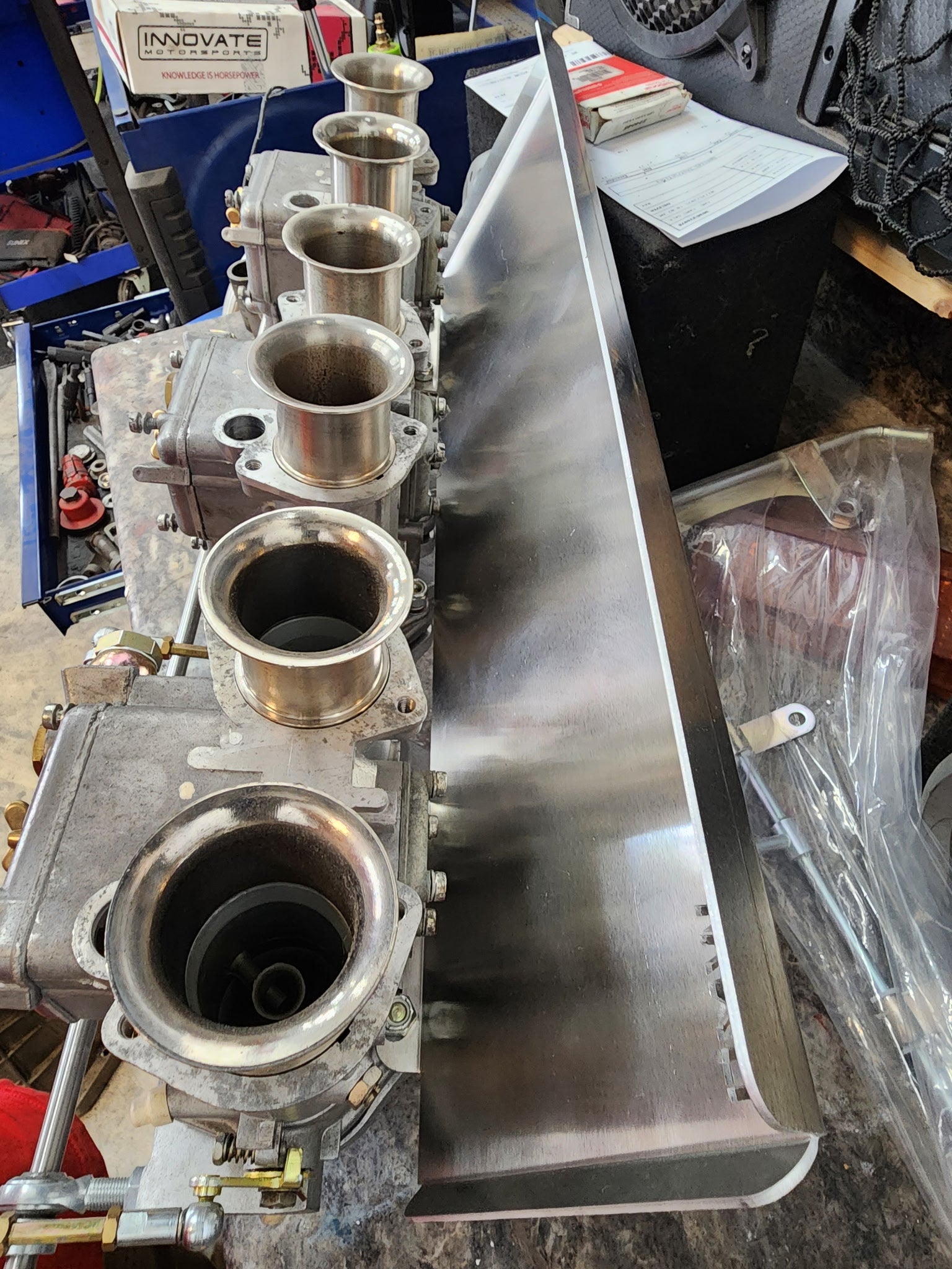 Row of automotive carburetors with velocity stacks and metal heat shield on workbench